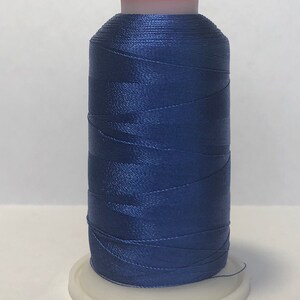 Puede incluir: Una bobina de hilo azul marino, bien enrollada y brillante, se encuentra sobre una base de pl&aacute;stico blanco. El hilo tiene un ligero brillo, y la parte superior de la bobina es de color rojo-naranja. Este hilo se utiliza para coser o bordar.