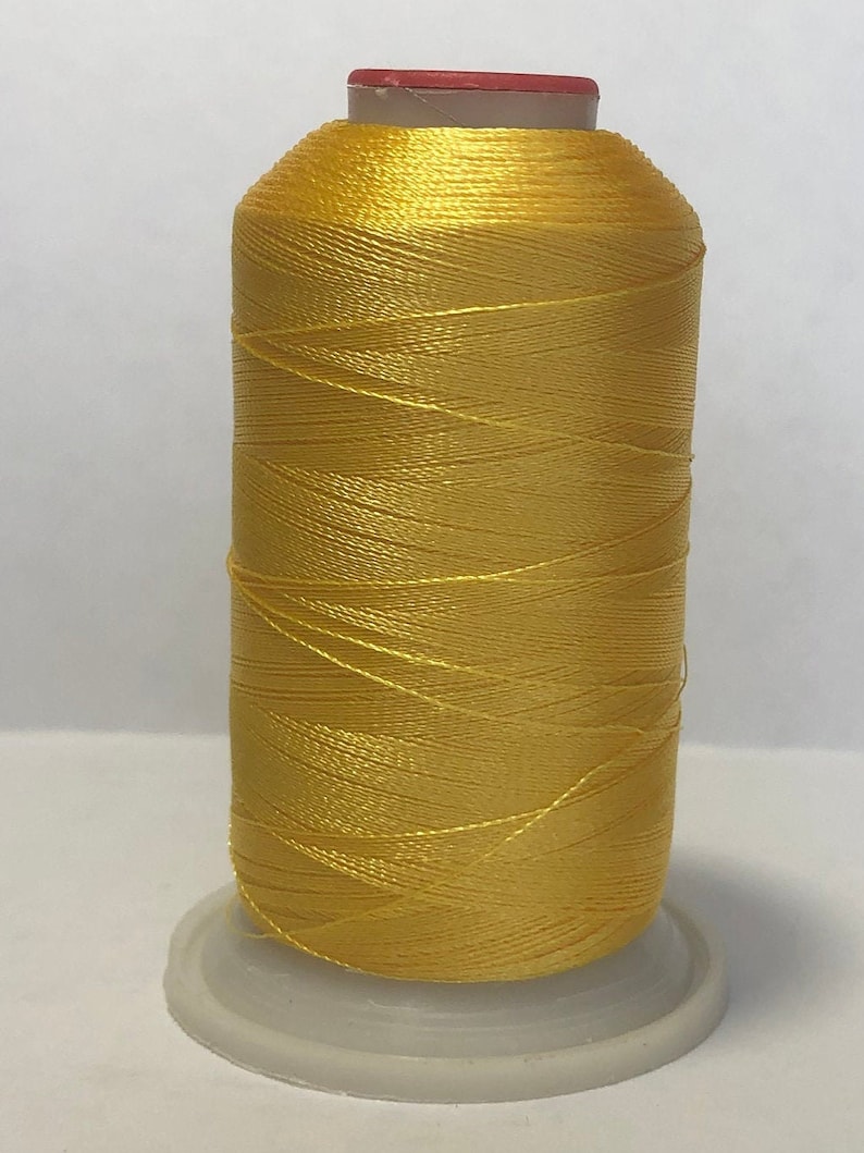 Puede incluir: Una bobina de hilo amarillo brillante, bien enrollada, con una parte superior roja y blanca. El hilo est&aacute; sobre una base de pl&aacute;stico blanco, adecuado para proyectos de costura y manualidades.