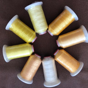 May include: An overhead shot displays eight spools of thread in a circular arrangement. The spools are various shades of yellow and gold, with white and red accents. The background is a solid brown.