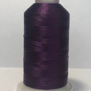 Puede incluir: Una bobina de hilo morado oscuro, con una base blanca y una parte superior roja. El hilo est&aacute; bien enrollado, creando una forma cil&iacute;ndrica. La imagen est&aacute; bien iluminada, resaltando la textura y el color del hilo.