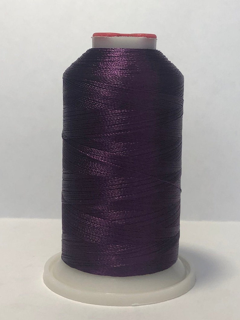 Puede incluir: Una bobina de hilo morado oscuro, con una base blanca y una parte superior roja. El hilo est&aacute; bien enrollado, creando una forma cil&iacute;ndrica. La imagen est&aacute; bien iluminada, resaltando la textura y el color del hilo.