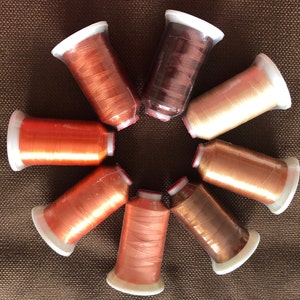 May include: A collection of eight spools of metallic thread in various shades of copper, bronze, and gold. Each spool is wrapped in clear plastic and has a white base. The threads are arranged in a circular pattern on a brown textured surface.