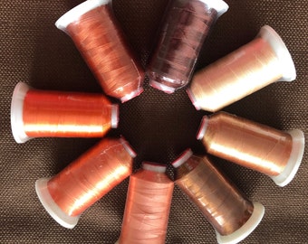 May include: A collection of eight spools of metallic thread in various shades of copper, bronze, and gold. Each spool is wrapped in clear plastic and has a white base. The threads are arranged in a circular pattern on a brown textured surface.