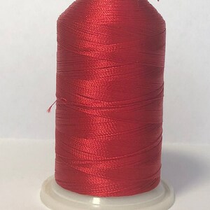 Puede incluir: Una bobina de hilo rojo vibrante, bien enrollado y brillante, se asienta sobre una base de pl&aacute;stico blanco. El hilo es de un color rico y saturado, ideal para coser, bordar u otras manualidades. La bobina tiene una parte superior roja y un peque&ntilde;o hilo que se extiende desde la parte superior.
