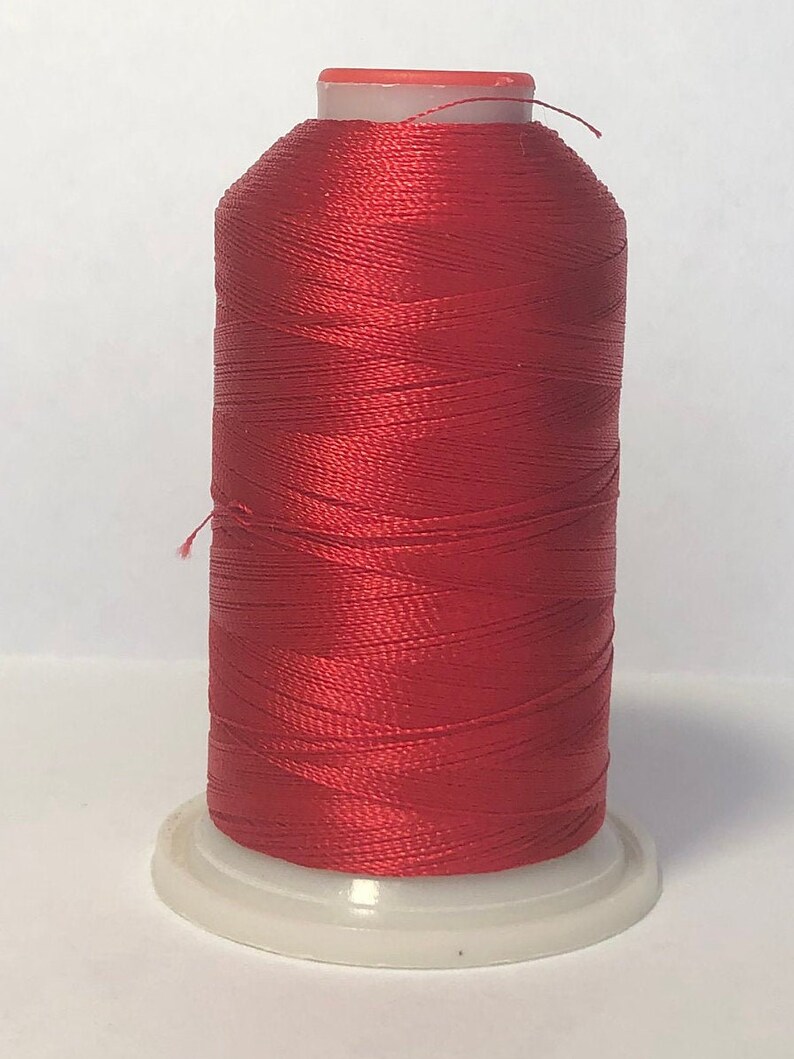 Puede incluir: Una bobina de hilo rojo vibrante, bien enrollado y brillante, se asienta sobre una base de pl&aacute;stico blanco. El hilo es de un color rico y saturado, ideal para coser, bordar u otras manualidades. La bobina tiene una parte superior roja y un peque&ntilde;o hilo que se extiende desde la parte superior.
