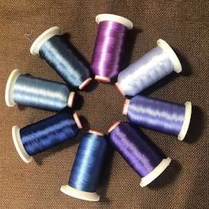 May include: An overhead shot of eight spools of thread in various shades of blue and purple. The spools are arranged in a circular pattern, with white bases and red accents. The thread appears to be a shiny, silky material.