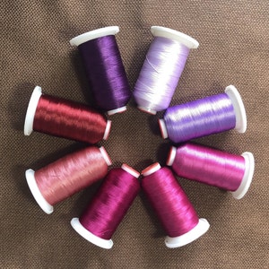 May include: An overhead shot displays eight spools of colorful thread arranged in a circular pattern. The threads are various shades of purple, pink, and red, with white spools. The background is a textured brown surface.