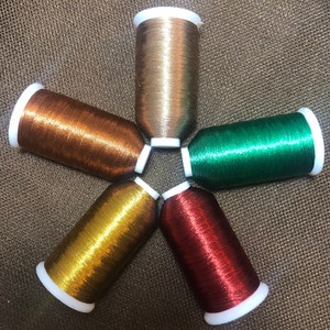 Puede incluir: Cinco carretes de hilo metálico en forma de estrella. Los colores incluyen dorado, bronce, verde, rojo y amarillo. Cada carrete tiene una base blanca y está lleno de hilo brillante y apretado. El fondo es una tela marrón texturizada.