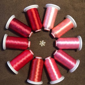 May include: Ten spools of embroidery thread arranged in a circle, featuring a gradient of colors from deep red to light pink. Each spool has white plastic ends. A small, silver-toned metallic snowflake ornament is positioned in the center.