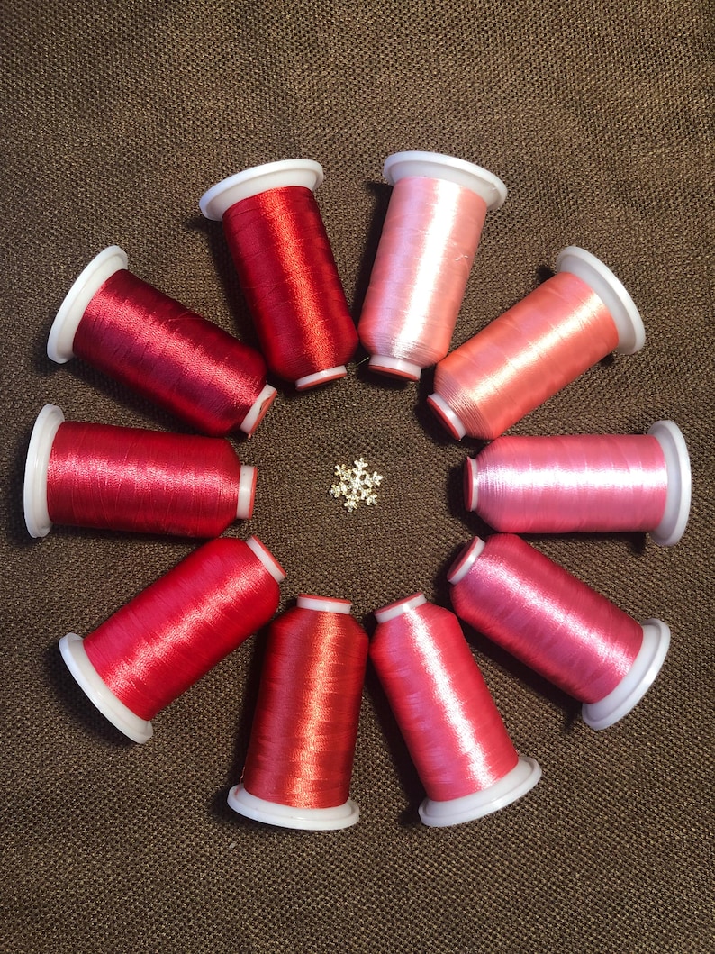 May include: Ten spools of embroidery thread arranged in a circle, featuring a gradient of colors from deep red to light pink. Each spool has white plastic ends. A small, silver-toned metallic snowflake ornament is positioned in the center.