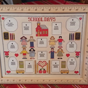 May include: A wooden picture frame with a ruler border and a cross-stitch design featuring a schoolhouse and 12 photo spaces for each grade of school. The text "SCHOOL DAYS" is at the top of the frame.