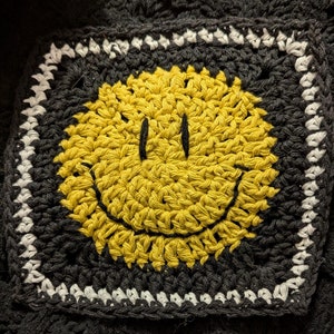 May include: A crocheted yellow smiley face with a black outline, framed by a black and white crocheted border.