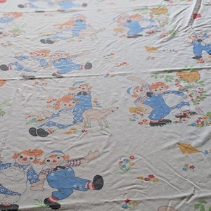 Może przedstawiać: Biała bawełniana tkanina z powtarzającym się wzorem lalek Raggedy Ann i Andy. Lalki są ubrane w niebieskie ogrodniczki i czerwono-białe kratkowane sukienki. Tło jest jasnoniebieskie z małymi białymi kwiatami.