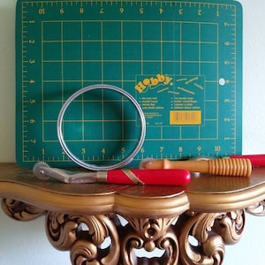May include: A set of three sewing tools on a gold shelf. The tools include a rotary cutter, a seam ripper, and a wooden tool with a red handle. The tools are on top of a green cutting mat with a ruler.