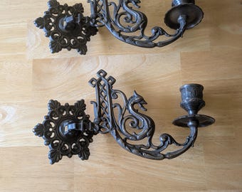 Könnte beinhalten: Zwei antike Wandleuchter aus Bronze. Jeder Leuchter hat ein verziertes Design mit einem Drachenmotiv und einem Kerzenhalter. Die Leuchter sind auf einer dekorativen, blütenförmigen Basis montiert. Das Gesamtdesign ist detailliert und vintage.
