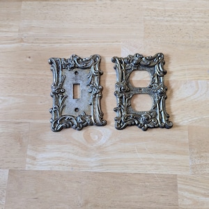 May include: Two ornate metal light switch covers with a decorative floral design. The cover on the left is for a single switch, and the cover on the right is for a double switch.