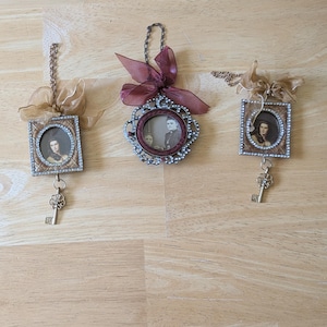 May include: Three antique-style pendants with miniature portraits. Two are square, one is round, all with ornate frames and decorative bows. Each pendant has a small key charm hanging below. The pendants are displayed on a light wood surface.