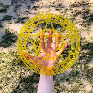 May include: A yellow plastic pentagram with a circle around it. The circle has various symbols and letters inscribed on it. The pentagram is held up by a hand.