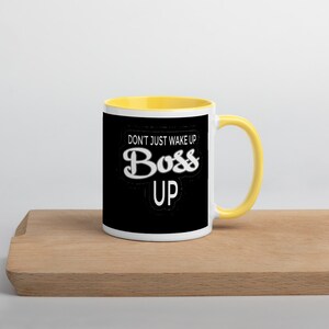 May include: A white ceramic mug with a yellow rim and handle. The mug has a black background with white text that reads "Don't just wake up Boss Up".