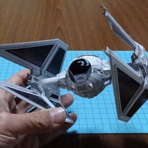 May include: A paper model of a Star Wars TIE fighter, predominantly light grey with black solar panels. The model has a hexagonal cockpit and detailed wings. The model is held in a person's hand, showcasing its intricate design.