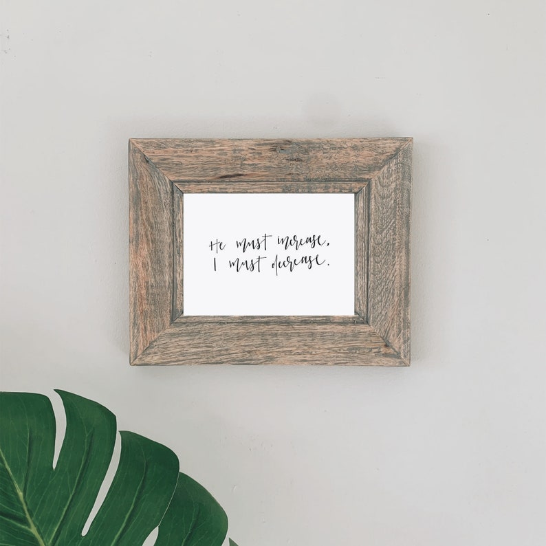 Pode incluir: Uma impress&atilde;o emoldurada com o texto manuscrito "Ele deve aumentar, eu devo diminuir". A moldura &eacute; feita de madeira desgastada e a impress&atilde;o est&aacute; em um fundo branco. A impress&atilde;o &eacute; exibida em uma parede cinza claro, com uma grande folha verde no canto inferior esquerdo.