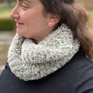 May include: A gray, fluffy infinity scarf made of a soft, textured material. The scarf is wrapped around the neck, creating a cozy and warm look. The background is blurred, with trees and foliage visible.