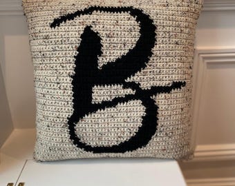 May include: A square crochet pillow featuring a speckled cream-colored yarn base with a large, bold, black letter B stitched in the center. The texture of the yarn is visible throughout the surface, showcasing the intricate loops of the crochet work.