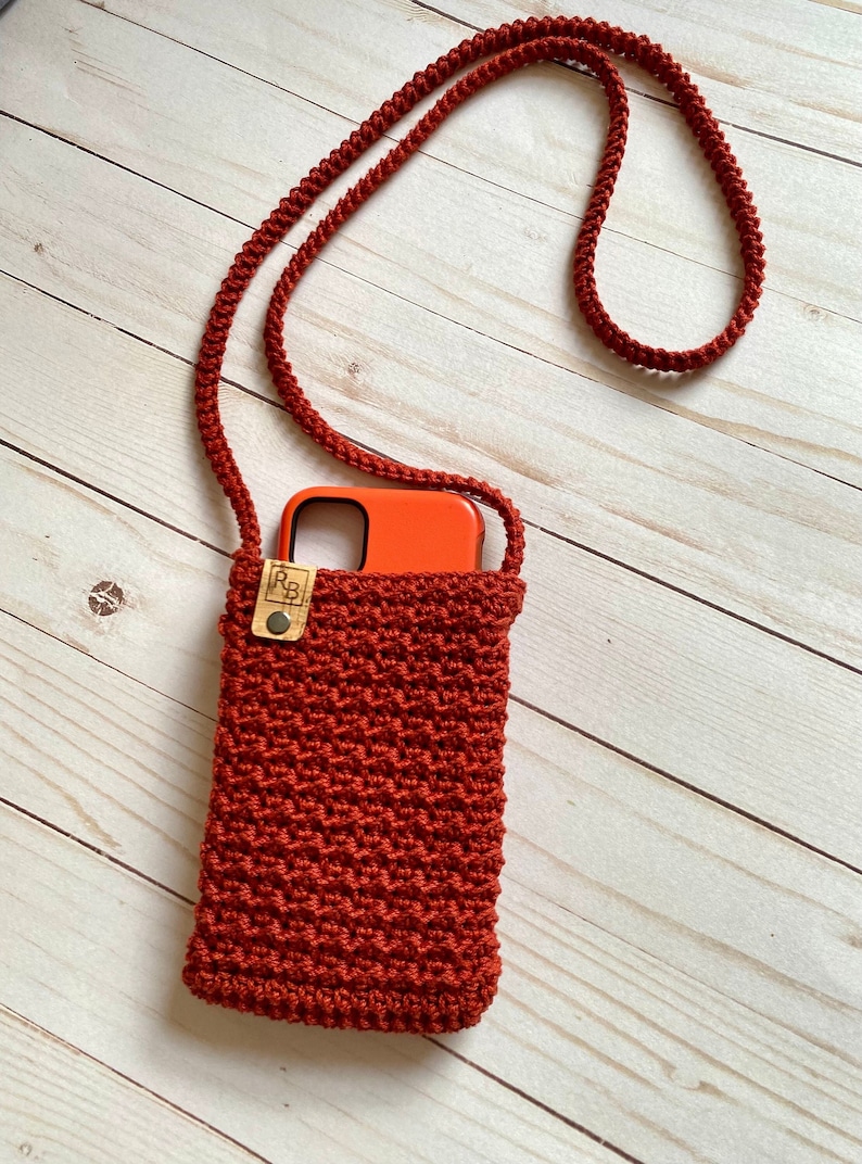 May include: A rust-colored, crocheted phone crossbody bag with a matching strap. The bag features a textured stitch pattern and a small wooden tag with a button closure. An orange phone is visible inside the bag.