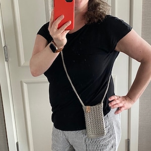 May include: A small, crocheted phone bag in a neutral color with a long strap. The bag has a rectangular shape and a woven texture. The strap is also crocheted. The bag is approximately 7 inches tall and 4 inches wide.