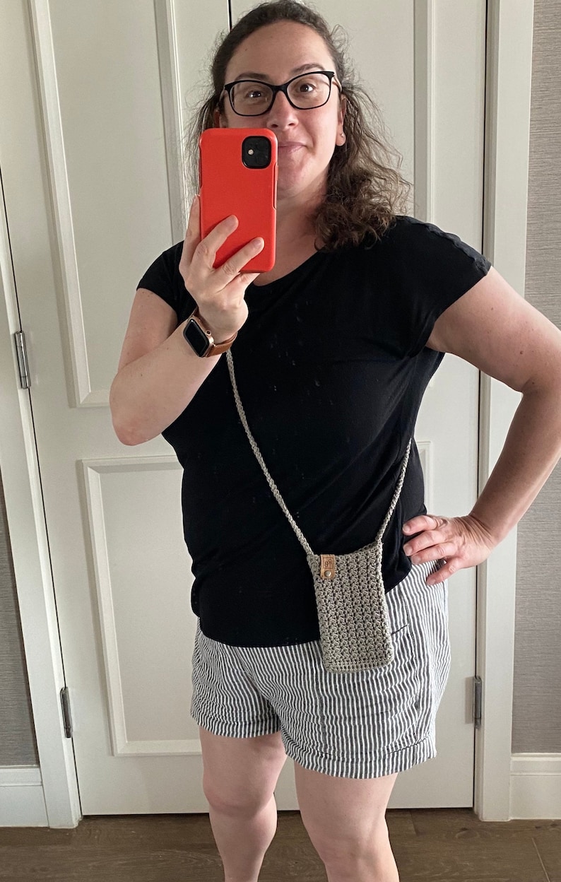 May include: A small, crocheted phone bag in a neutral color with a long strap. The bag has a rectangular shape and a woven texture. The strap is also crocheted. The bag is approximately 7 inches tall and 4 inches wide.