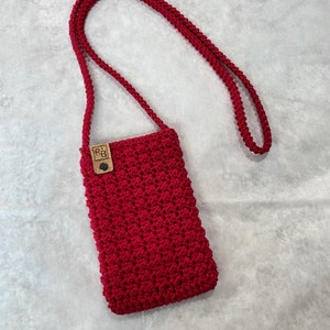 May include: A handmade, red macrame phone crossbody bag. The bag features a textured, woven design and a long, braided strap. A small, wooden tag with the initials "R&B" is attached to the front of the bag.
