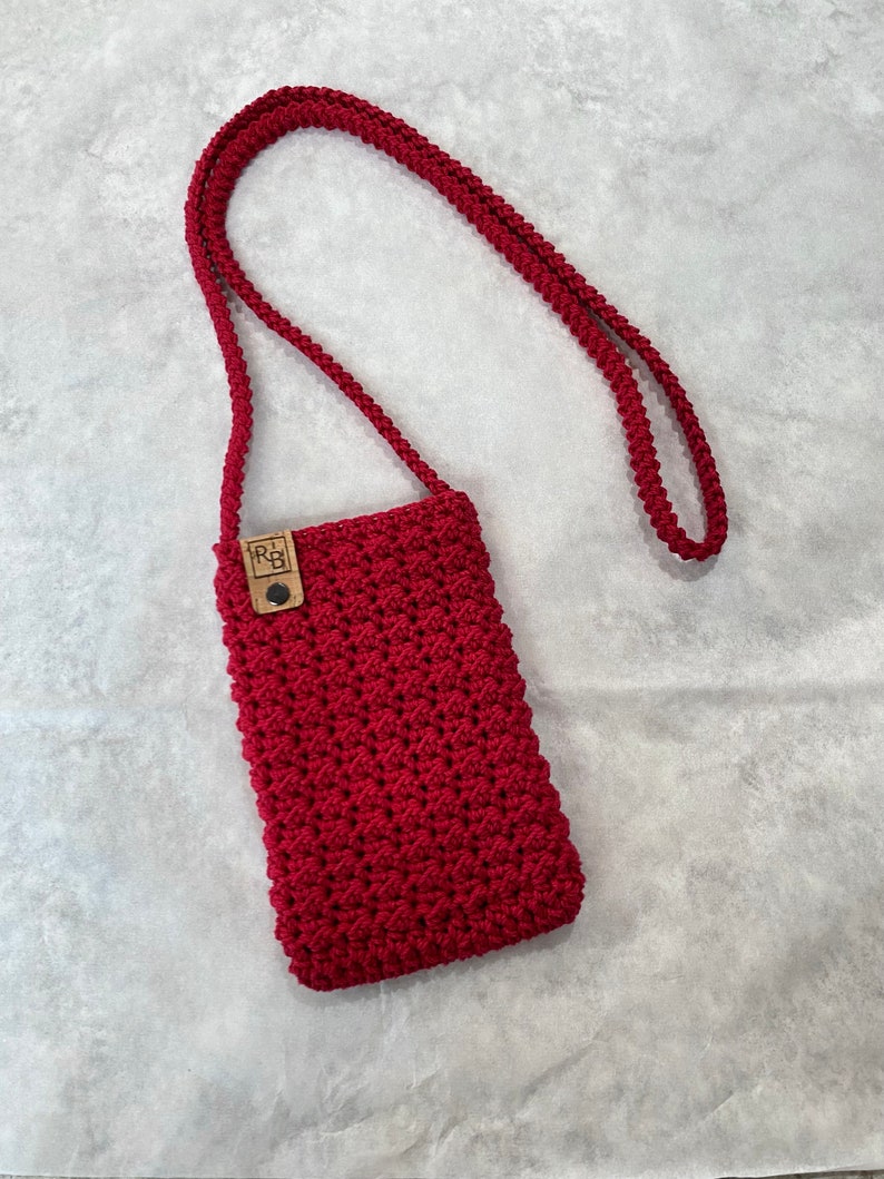May include: A handmade, red macrame phone crossbody bag. The bag features a textured, woven design and a long, braided strap. A small, wooden tag with the initials "R&B" is attached to the front of the bag.