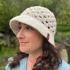 May include: A cream-colored crochet bucket hat with a small brim. The hat features an open weave design and a small brown leather tag. The hat is made of a soft, textured yarn, perfect for sun protection.