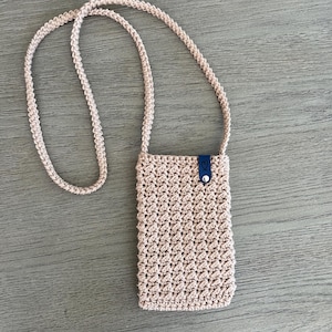 May include: A light beige, crocheted crossbody phone bag with a long strap. The bag has a textured pattern and a small, dark blue leather tab with a button closure. The strap is also crocheted in the same beige color.