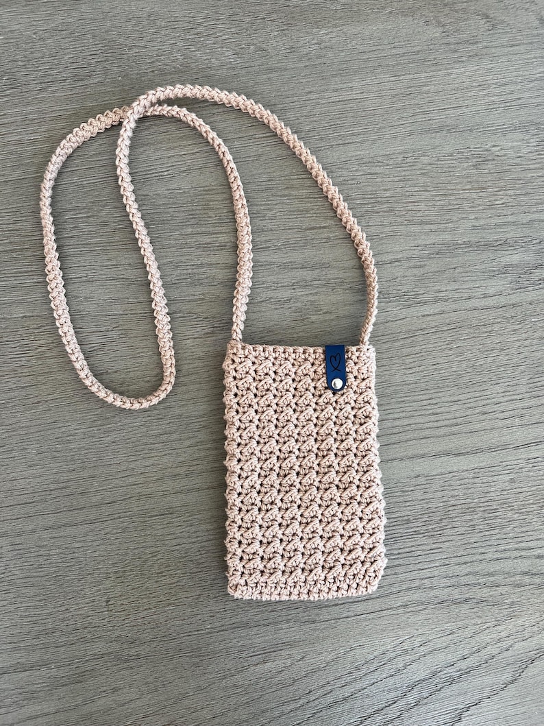 May include: A light beige, crocheted crossbody phone bag with a long strap. The bag has a textured pattern and a small, dark blue leather tab with a button closure. The strap is also crocheted in the same beige color.