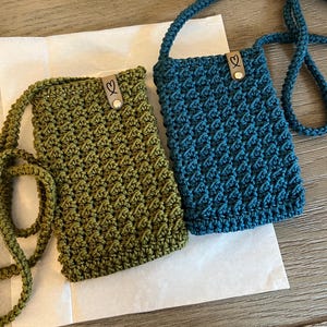 May include: Two handmade crochet phone bags in olive green and teal blue. Each bag features a textured stitch pattern, a long strap, and a small leather tag with a heart detail. The bags are displayed on a white paper surface.