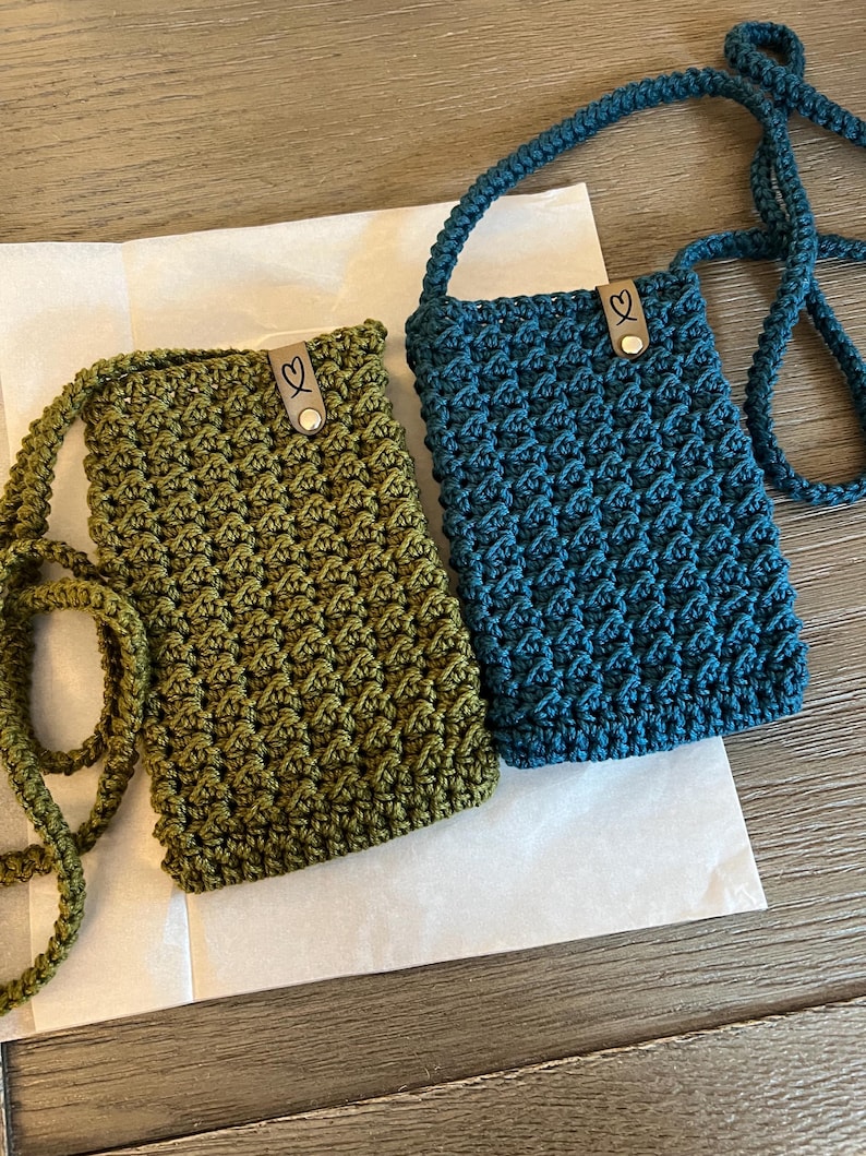 May include: Two handmade crochet phone bags in olive green and teal blue. Each bag features a textured stitch pattern, a long strap, and a small leather tag with a heart detail. The bags are displayed on a white paper surface.