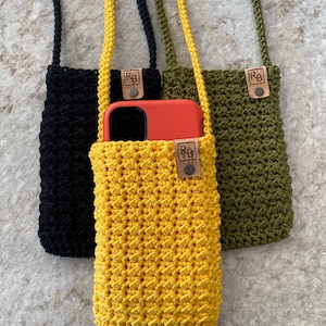 May include: Three handmade crochet phone crossbody bags in black, yellow, and olive green. Each bag has a long strap and a leather tag with the initials "RB". The yellow bag holds an orange phone.