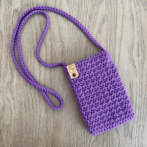 May include: A handmade, purple crochet phone crossbody bag with a matching strap. The bag features a textured stitch pattern and a small, rectangular wooden tag with the initials "RB". The bag is shown on a wooden surface.