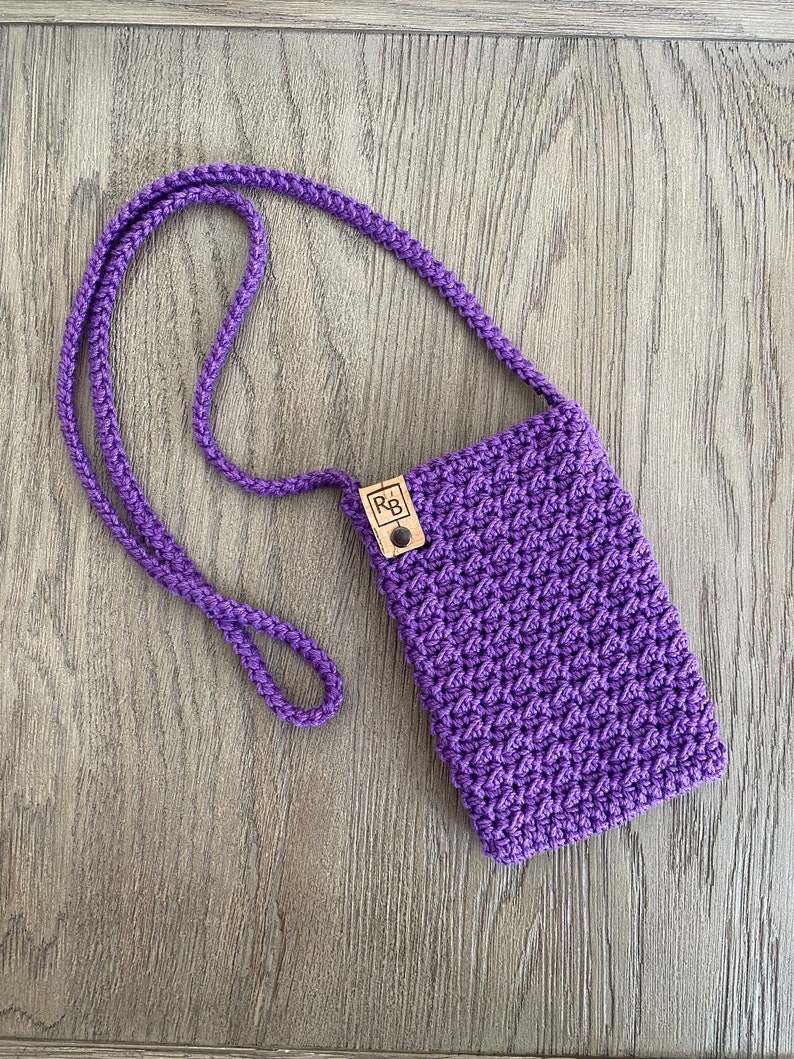 May include: A handmade, purple crochet phone crossbody bag with a matching strap. The bag features a textured stitch pattern and a small, rectangular wooden tag with the initials "RB". The bag is shown on a wooden surface.