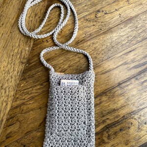 May include: A light gray, crocheted phone crossbody bag with a matching strap. The bag has a pocket with a card inside. The bag is made of a textured yarn and is designed to be worn around the neck.