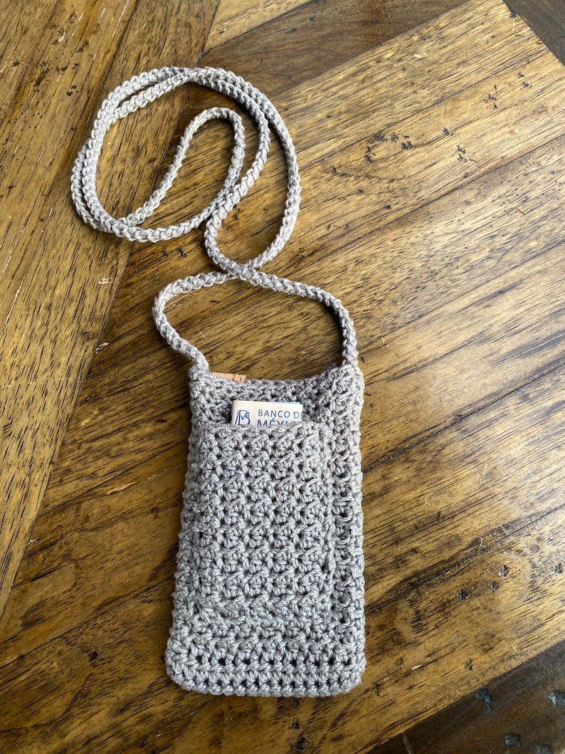 May include: A light gray, crocheted phone crossbody bag with a matching strap. The bag has a pocket with a card inside. The bag is made of a textured yarn and is designed to be worn around the neck.