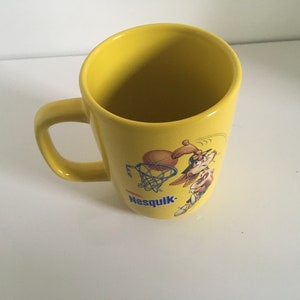 Peut inclure: Une tasse en céramique jaune avec un lapin de dessin animé jouant au basket-ball et le texte "Nesquik" imprimé sur le côté.