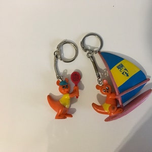 May include: Two orange kangaroo keychains. One is holding a red tennis racket and the other is on a pink surfboard with a blue and yellow sail.
