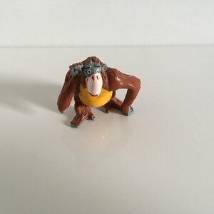 May include: A brown and gray plastic figurine of an orangutan wearing a yellow shirt. The orangutan is holding its hand to its ear.