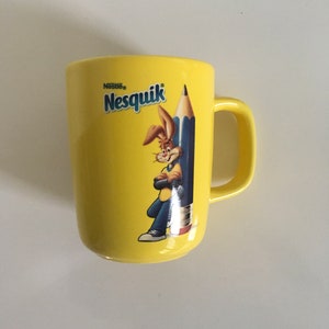 Peut inclure: Une tasse en céramique jaune avec le logo Nesquik et un lapin de dessin animé tenant un crayon. Le lapin porte une chemise bleue et des chaussures rouges.