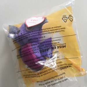 Op de afbeelding: Een paars en zilveren pluche speelgoed met een hartvormig label waarop "Levi" staat. Het speelgoed is verpakt in een doorzichtige plastic zak met een gele achtergrond. De zak bevat een waarschuwing over verstikkingsgevaar en instructies voor het weggooien.