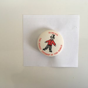 May include: A white round button with red text that reads "Hamleys The Finest Toyshop In The World". A black and white illustration of a person wearing a red jacket and a top hat is in the center of the button.