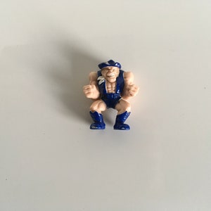Puede incluir: Una pequeña figura de acción de plástico de un luchador que lleva un atuendo azul y blanco. La figura tiene una barba blanca y un sombrero azul.