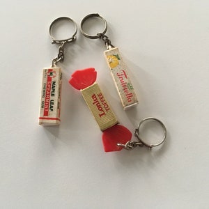May include: Three vintage keychains featuring candy wrappers. The keychains are made of metal and have red and white candy wrappers with the words "Toffee Lollies", "Maple Leaf Spearmint Chewing Gum", and "Fruittella" printed on them.